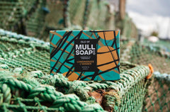 a lemongrass and ginger soap box sitting on a green creel pot on Tobermory pier. The box design is green with yellow buoys and black fishing nets. The soap is described as warm and spicy. Handmade in Tobermory by the Isle of Mull soap co.  