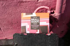 a Scottish rose and patchouli soap box  displayed next to a sunny wall painted pink. The box is a pink colourful design. The soap is described as mossy, earthy and floral. Handmade in Tobermory by the Isle of Mull soap co.  