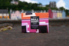 a Scottish rose and patchouli soap box  displayed on Tobermory pier. The box is a pink colourful design shown infront of the colourful Main Street of houses and shops.. The soap is described as mossy, earthy and floral. Handmade in Tobermory by the Isle of Mull soap co.  