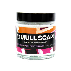 A glass jar with black lid. colourful pink label design, filled with Scottish rose and patchouli hand and body cream handmade in Tobermory by the Isle of Mull soap co. 